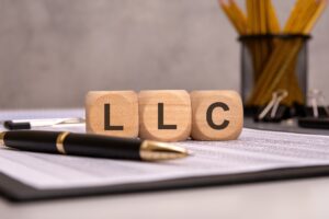 Three wooden letter blocks spelling "LLC" sit on a paper on a clipboard next to a fountain pen.