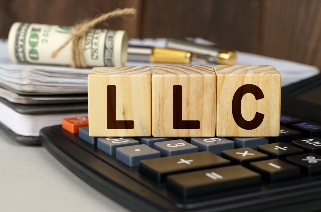 Calculator with blocks ontop that say LLC infront of rolled up money and paperwork