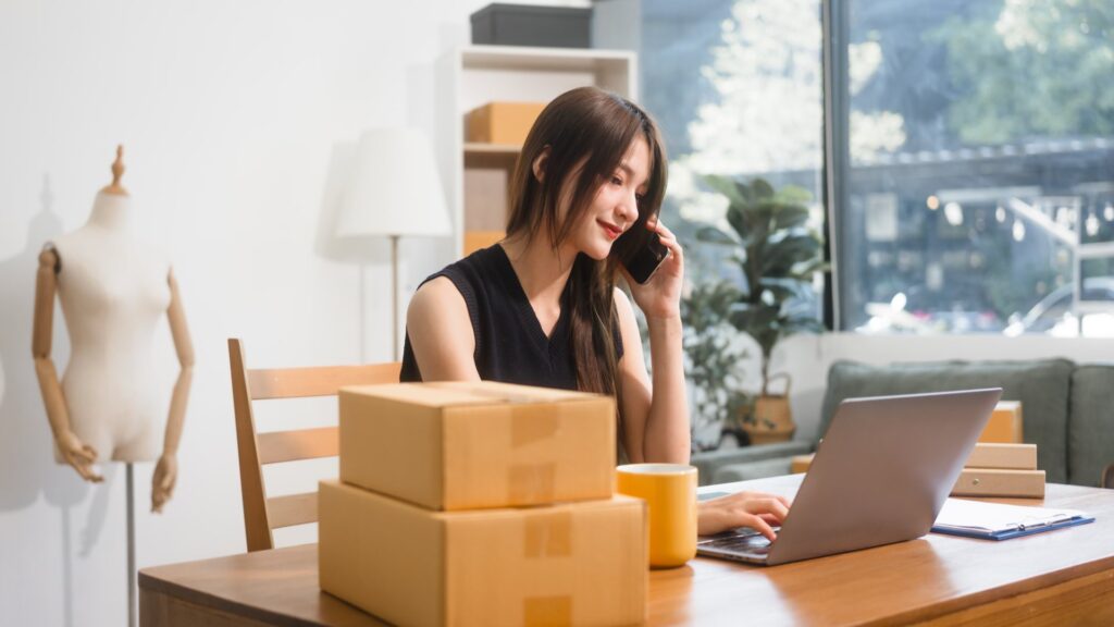 Woman sitting at a desk on the phone typing on a laptop next to boxes