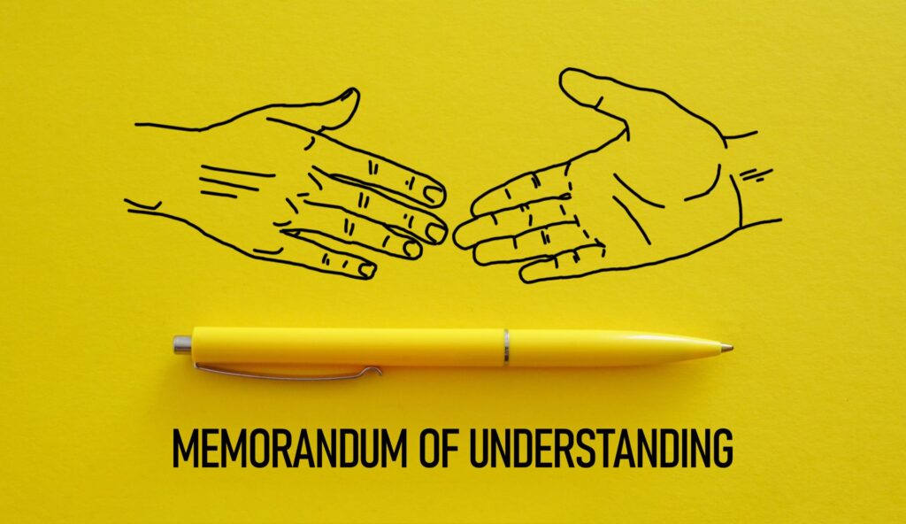 Two hands getting ready to shake a yellow pen and under it says Memorandum of Understanding