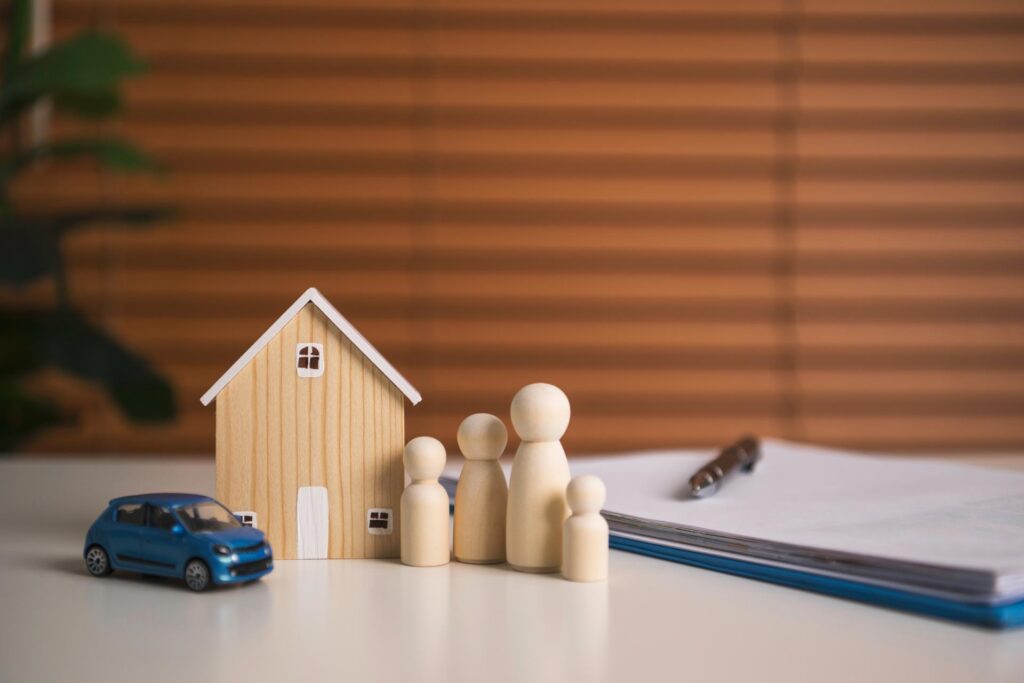 Wooden models of a family and a house sit next to a toy car and paperwork on a clipboard with a pen.