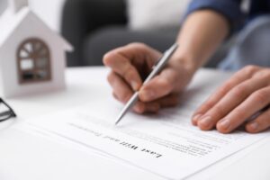 A person holding a silver pen writes on an estate planning document next to a wooden model of a house.